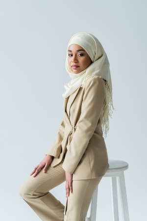 stylish muslim woman in hijab and beige suit leaning on stool isolated on greyの写真素材