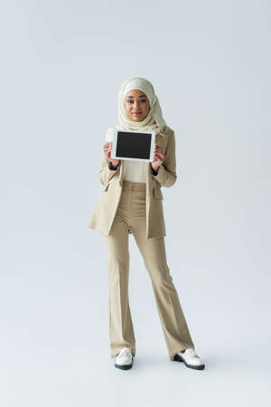 full length of pleased muslim woman in hijab and stylish suit holding digital tablet with blank screen on greyの写真素材