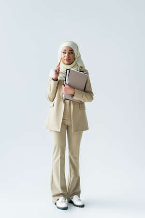 full length of pensive muslim woman in hijab holding pencil and folders on greyの写真素材