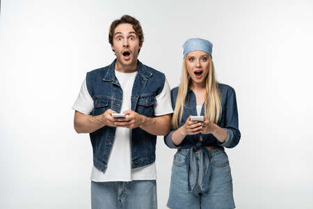 astonished couple in trendy denim clothes looking at camera while using cellphones isolated on whiteの写真素材