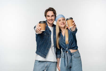 smiling couple in trendy denim clothes showing paper cups isolated on white