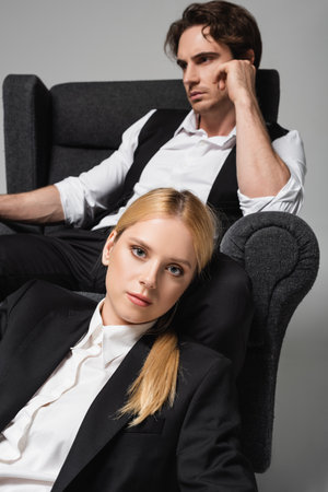 elegant blonde woman looking at camera near blurred man sitting in armchair isolated on greyの写真素材