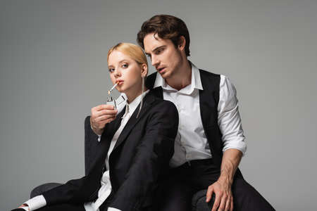 stylish man in elegant clothes lightening cigarette of blonde woman isolated on grey
