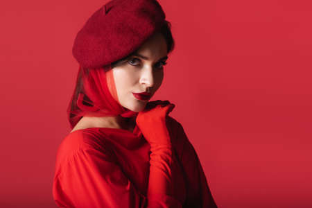 elegant young woman in beret and glove looking at camera isolated on redの写真素材