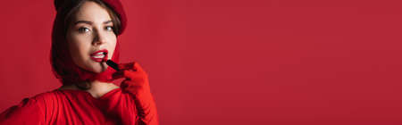 young woman in glove, headscarf and beret applying lipstick isolated on red, bannerの写真素材