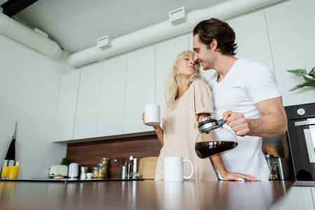 happy man holding coffee pot near blonde girlfriend in kitchenの写真素材
