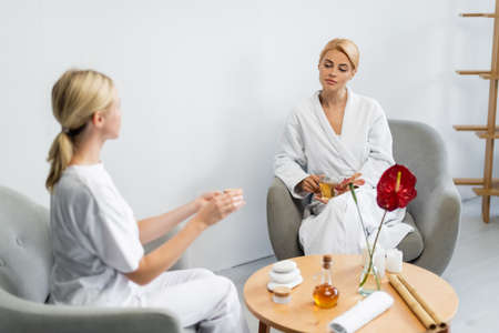 blonde client in bathrobe holding cup of tea while looking at blurred spa specialist with cream containerの写真素材