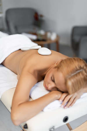 blurred and blonde woman with closed eyes receiving hot stone massage in spa centerの写真素材