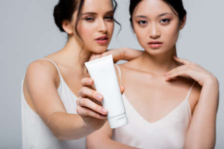 Blurred woman holding cosmetic cream near asian friend isolated on greyの写真素材