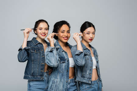Cheerful interracial women in denim jackets holding cosmetic brushes near eyes isolated on greyの写真素材
