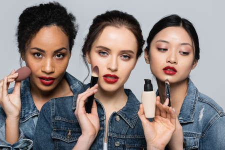 Interracial women with red lips holding cosmetic brushes and face foundation isolated on greyの写真素材