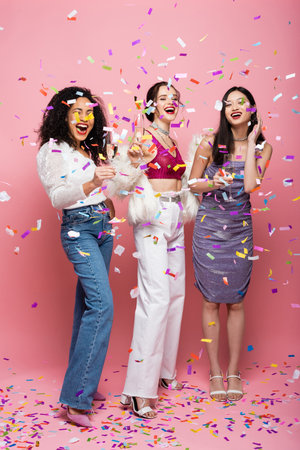 Pretty multicultural women with champagne looking at camera under confetti on pink backgroundの写真素材