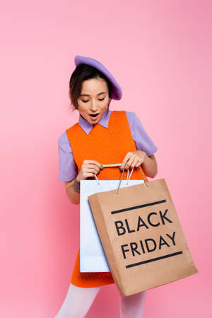 amazed woman in purple beret looking at shopping bag with black friday lettering isolated on pinkの写真素材