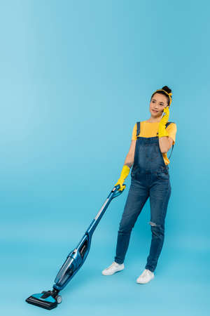 woman in denim overalls and rubber gloves talking on smartphone while vacuuming on blueの写真素材