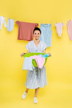 full length view of smiling housewife holding laundry bowl near clothes hanging on yellow backgroundの写真素材