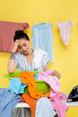 exhausted housewife touching head while standing near laundry on ironing board on yellowの写真素材