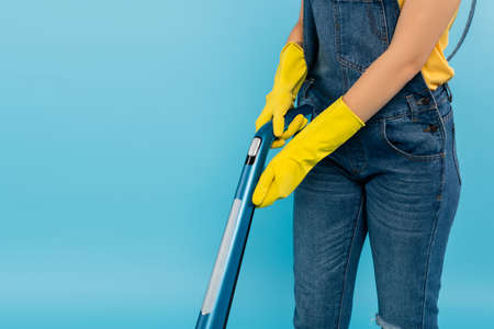 partial view of housewife in denim overalls and yellow rubber gloves vacuuming isolated on blue
