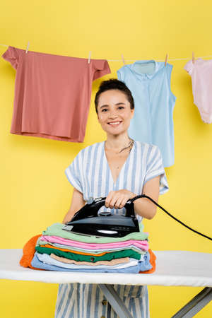 pleased woman holding iron near stack of clothes on ironing board on yellowの写真素材