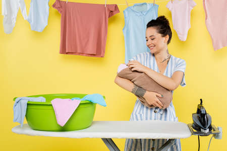 smiling woman holding baby doll near laundry bowl on ironing board on yellowの写真素材
