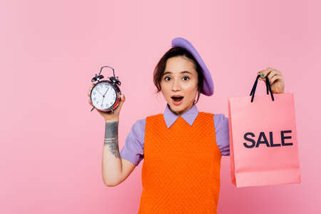 excited woman in bright clothes holding vintage alarm clock and sale shopping bag isolated on pinkの写真素材