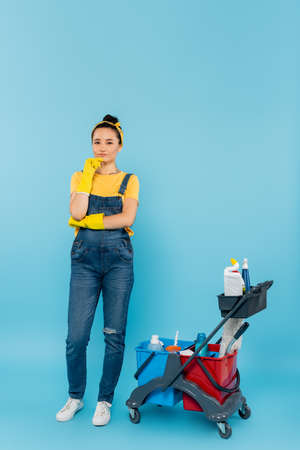 cleaner in rubber gloves and denim overalls looking at camera near cart with cleaning supplies on blueの写真素材