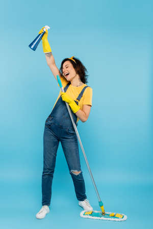 excited woman in rubber gloves and denim overalls singing with detergent and mop on blueの写真素材