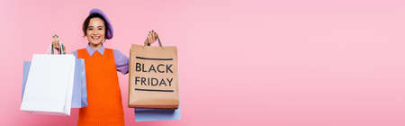 smiling woman holding purchases and shopping bag with black friday lettering isolated on pink,...