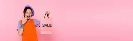 astonished woman with sale shopping bag covering open mouth with hand isolated on pink, bannerの写真素材
