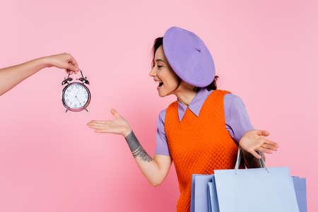 amazed woman showing wow gesture near female hand with vintage alarm clock isolated on pinkの写真素材