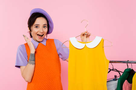 amazed woman showing wow gesture near yellow sleeveless dress and rack with clothes isolated on pink