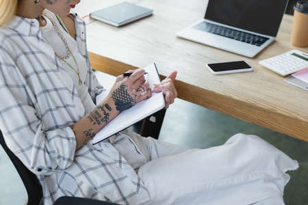 cropped view of tattooed businesswoman writing in notebook near gadgets with blank screen on deskの写真素材