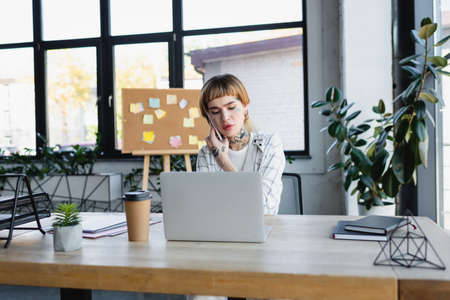 tattooed manager talking on mobile phone near laptop in modern office