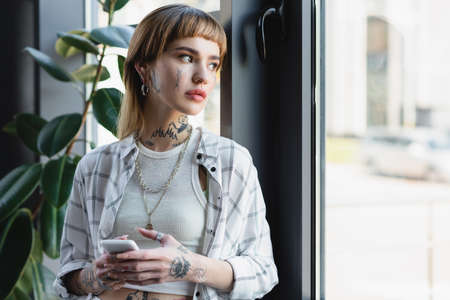 young tattooed woman with mobile phone standing near window in officeの写真素材
