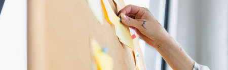 cropped view of woman with tattooed hand attaching sticky note on corkboard, blurred foreground,...