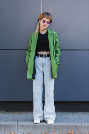 tattooed woman in jeans standing with hands in pockets of green jacket near grey wall