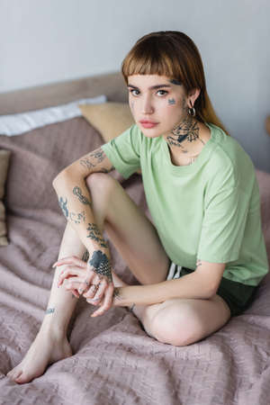young tattooed woman sitting on bed at home and looking at cameraの写真素材