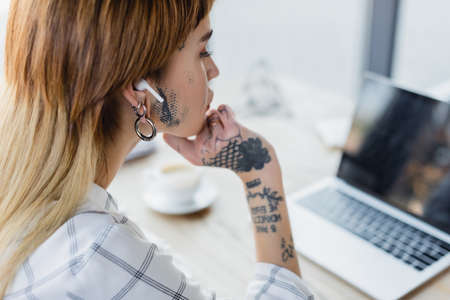 tattooed businesswoman in wireless earphone looking at blurred laptop in officeの写真素材