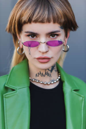 stylish tattooed woman in eyeglasses looking at camera outdoors