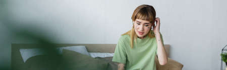 tattooed woman touching head while sitting in bedroom on blurred foreground, banner