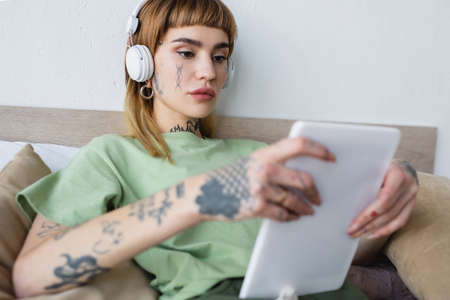 young tattooed woman with blurred digital tablet listening podcast in bedroomの写真素材