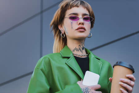 low angle view of tattooed woman with coffee to go and smartphone looking at camera outdoors