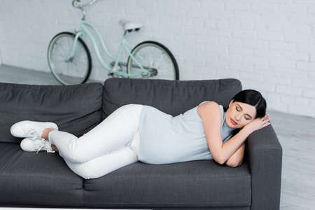 full length view of young pregnant woman sleeping on couch near blurred bike in living roomの写真素材