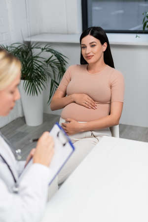 happy pregnant woman looking at blurred doctor writing prescription on clipboardの写真素材