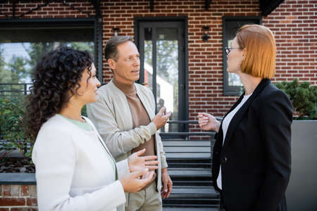 redhead realtor in glasses talking with multiethnic couple near modern house