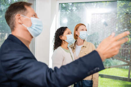 blurred realtor in medical mask pointing with hand near interracial lesbian couple inside of new houseの写真素材