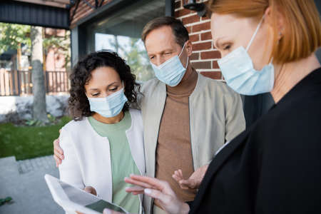blurred realtor in medical mask showing digital tablet to interracial couple near new house