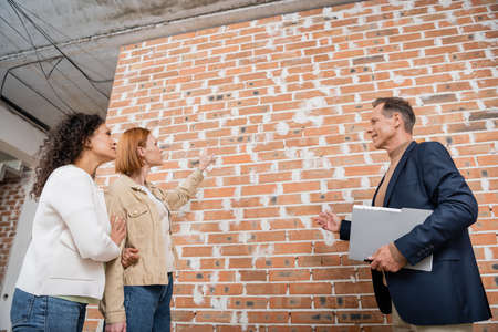 realtor with folder pointing at brick wall near interracial lesbian couple