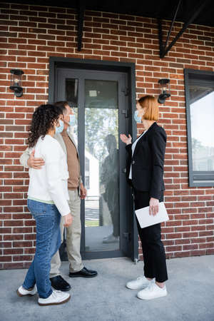 redhead realtor in medical mask holding digital tablet and pointing at new house near interracial coupleの写真素材