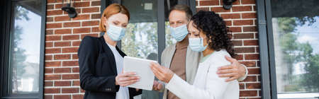 redhead realtor in medical mask showing digital tablet to interracial couple near new house, banner