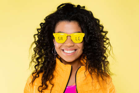 smiling african american young woman in sunglasses with sale lettering isolated on yellow